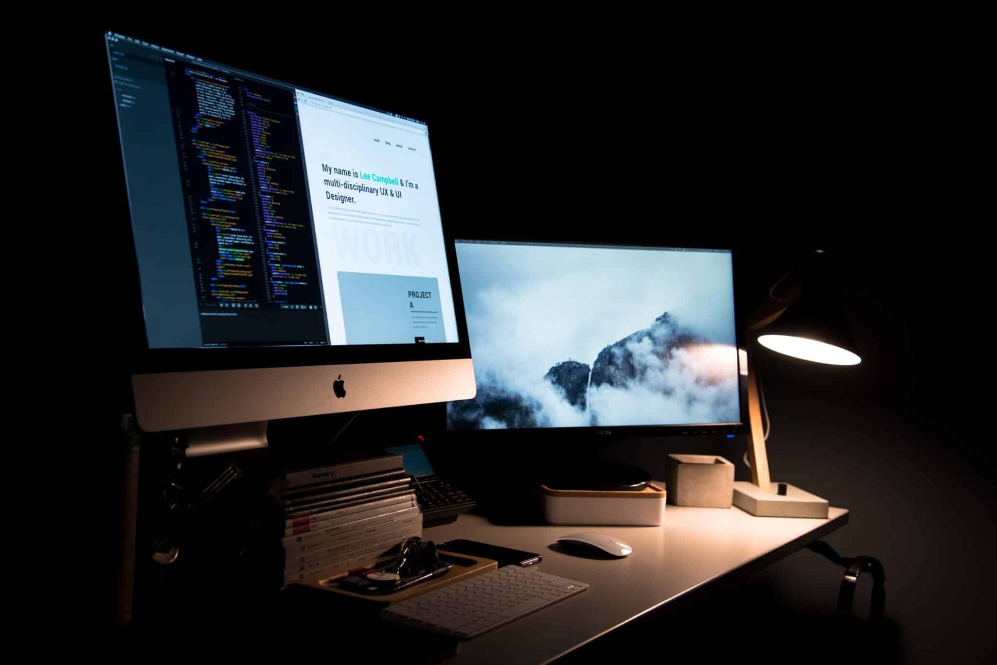 Two monitors on a desk in a darkened room, one showing code and one showing a website design.