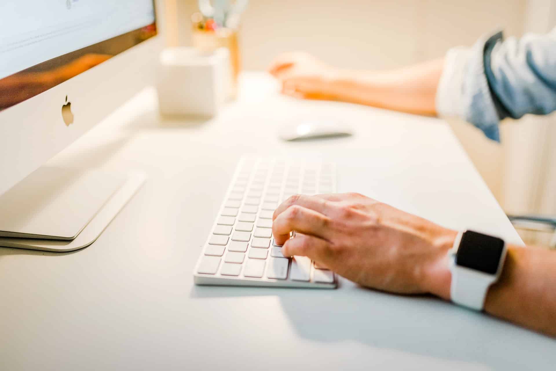 A person typing on a keyboard at a bright desk with an iMac and a smartwatch on their wrist.