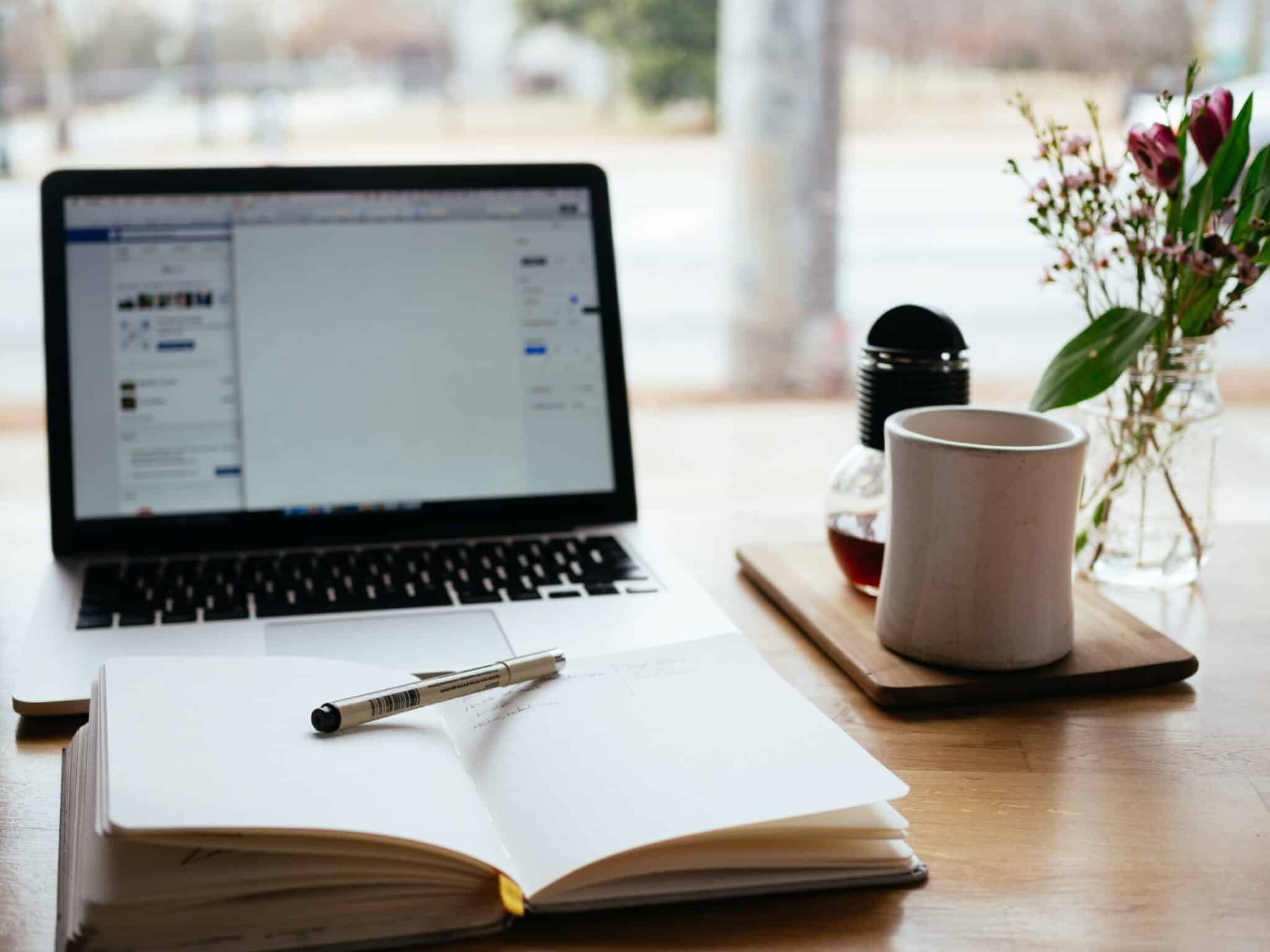 A laptop open on a cafe table beside an open notebook, a pen, a cup of coffee and a vase of flowers.