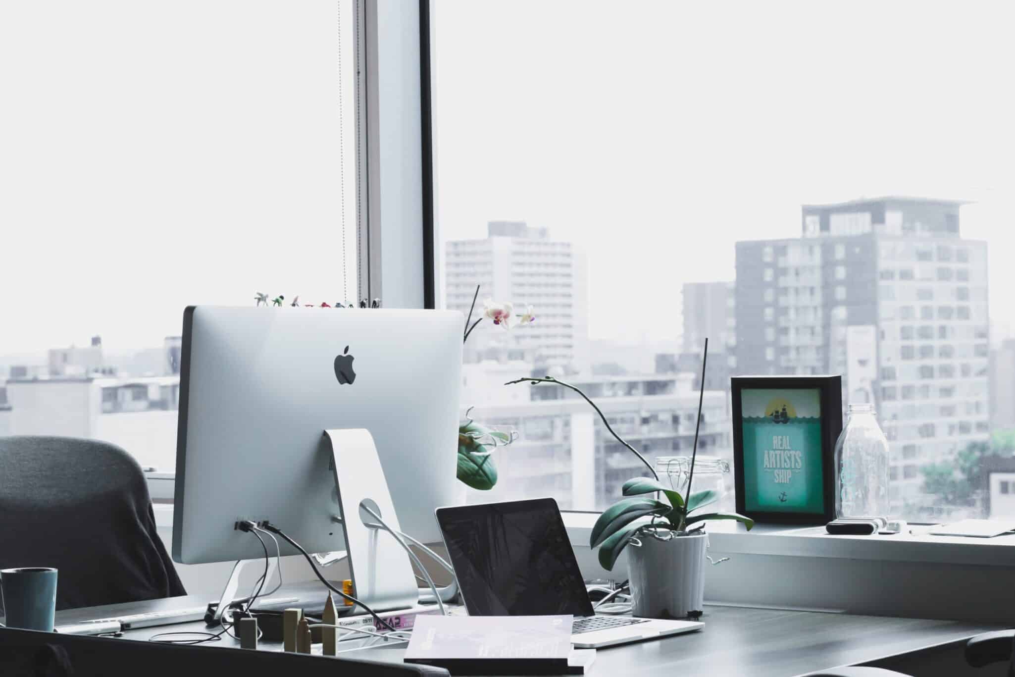 An iMac on a desk beside a window overlooking a city skyline.