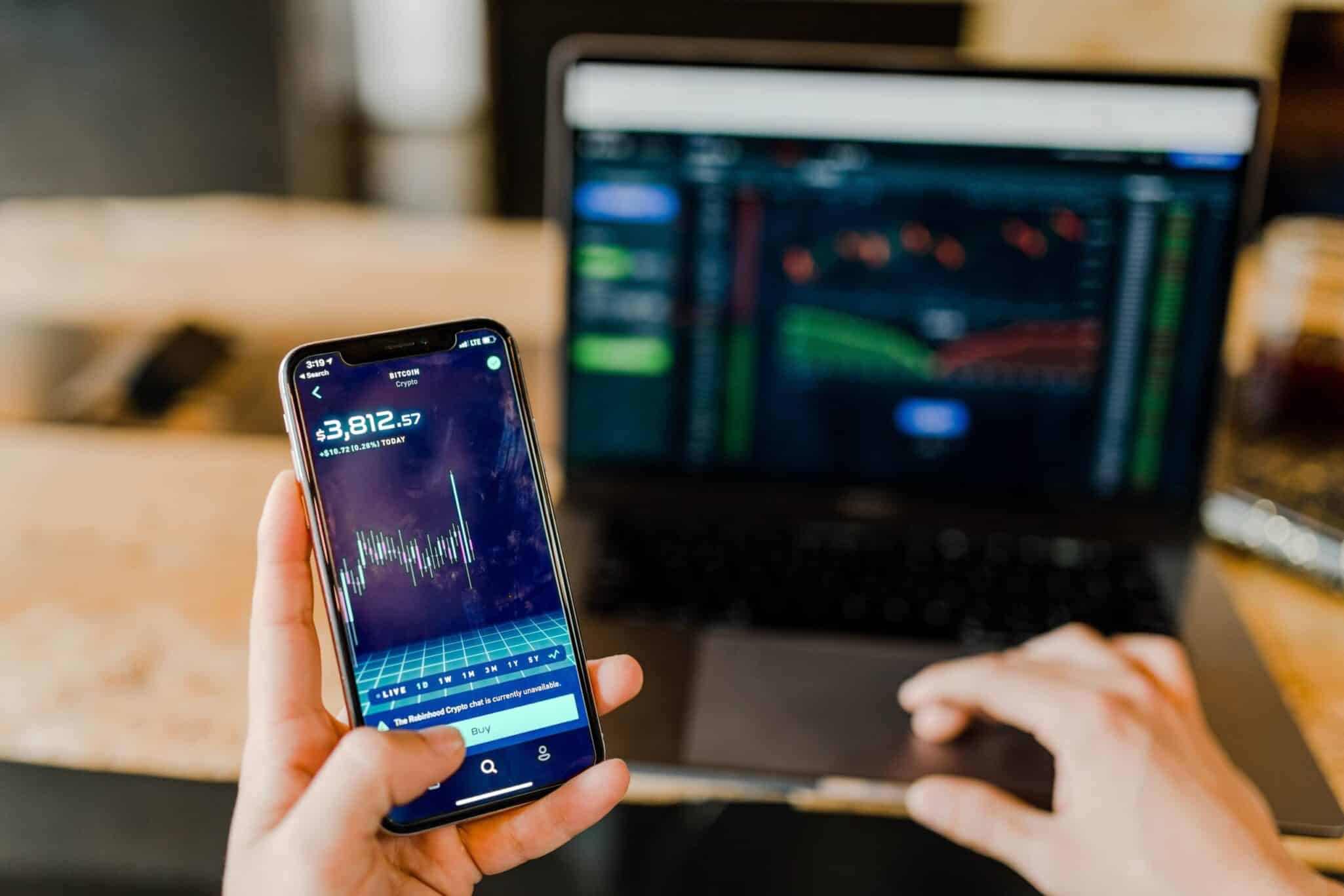 A hand holding a phone showing a stock trading app, with a laptop displaying trading charts behind it.