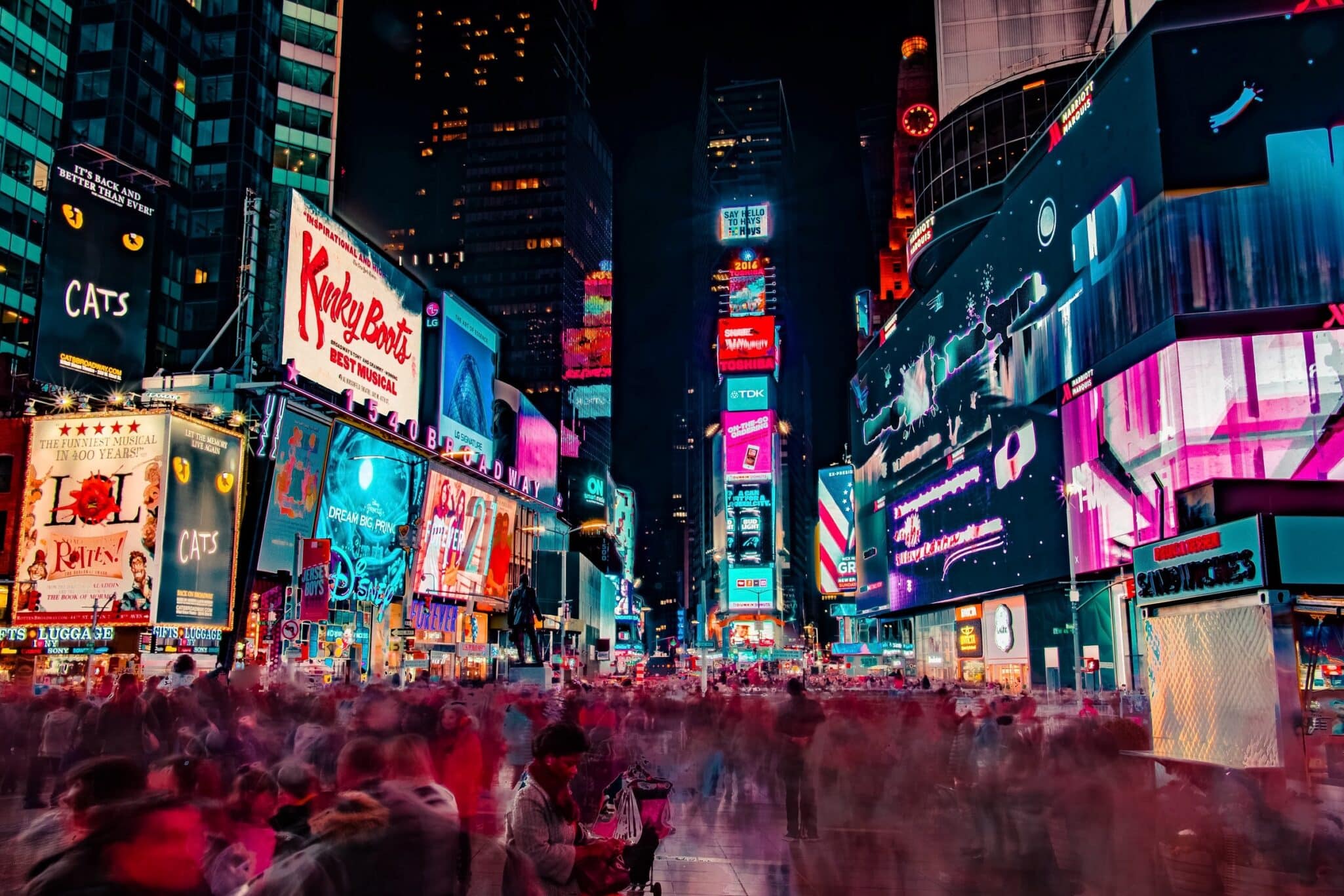 Times Square at night, crowded with people beneath a wall of illuminated advertising screens.