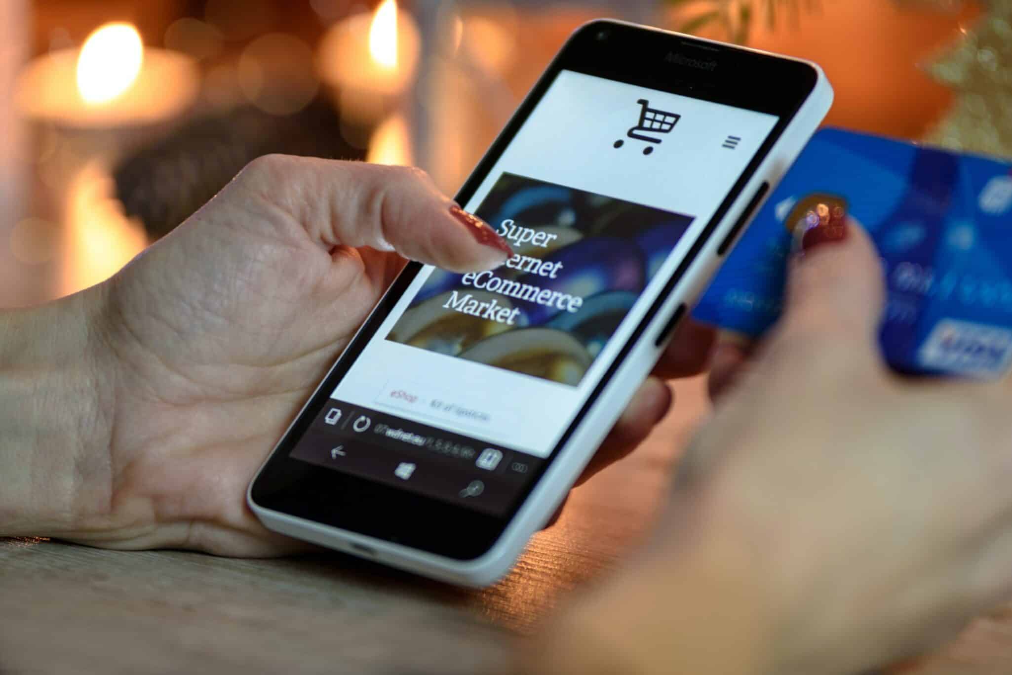 A hand holding a phone showing an ecommerce app, with a bank card held in the other hand.