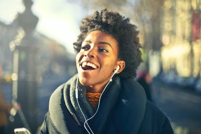 A woman laughing while listening to music through earphones on a sunny street.