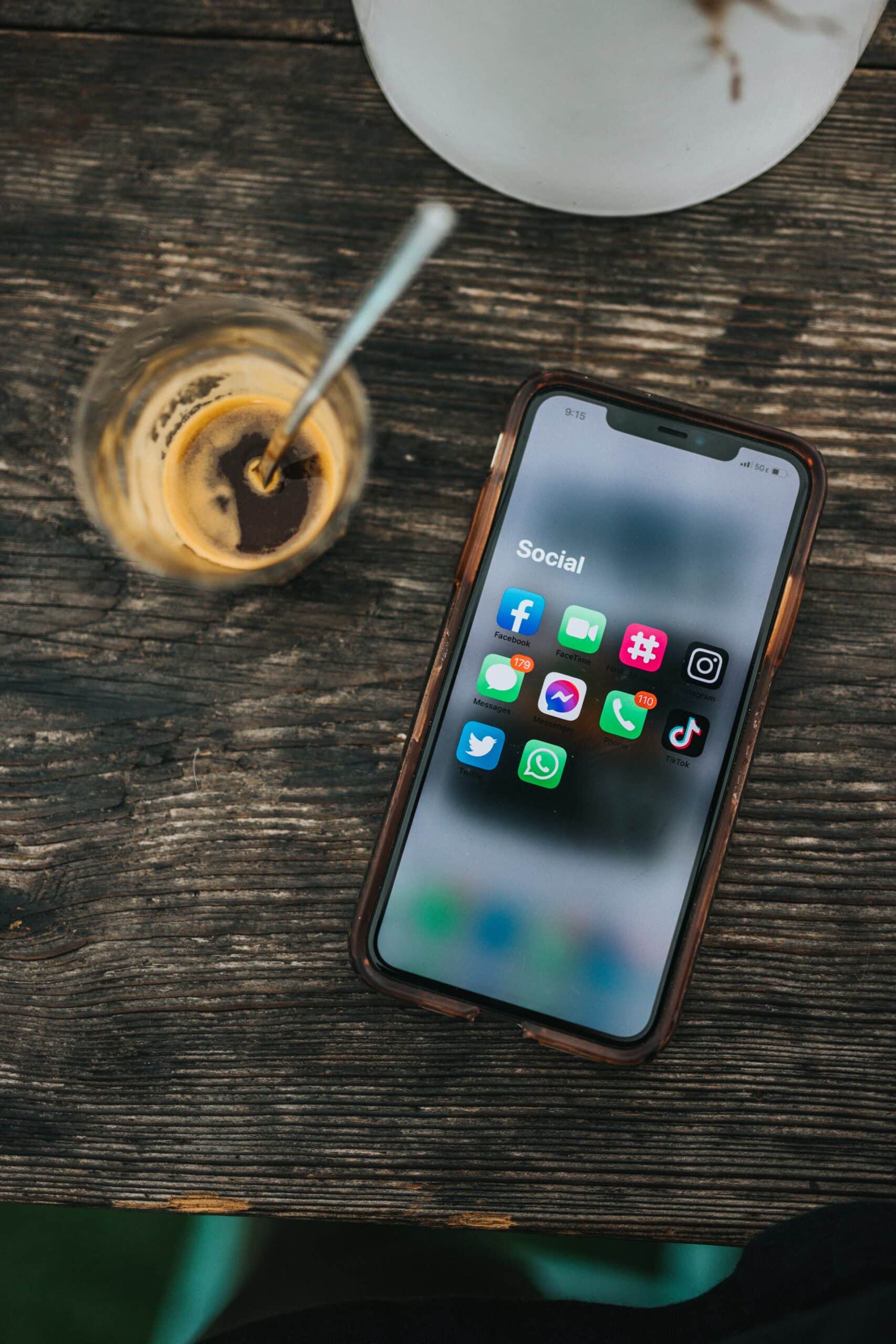 A phone on a wooden table showing a folder of social media apps, beside a glass of iced coffee.