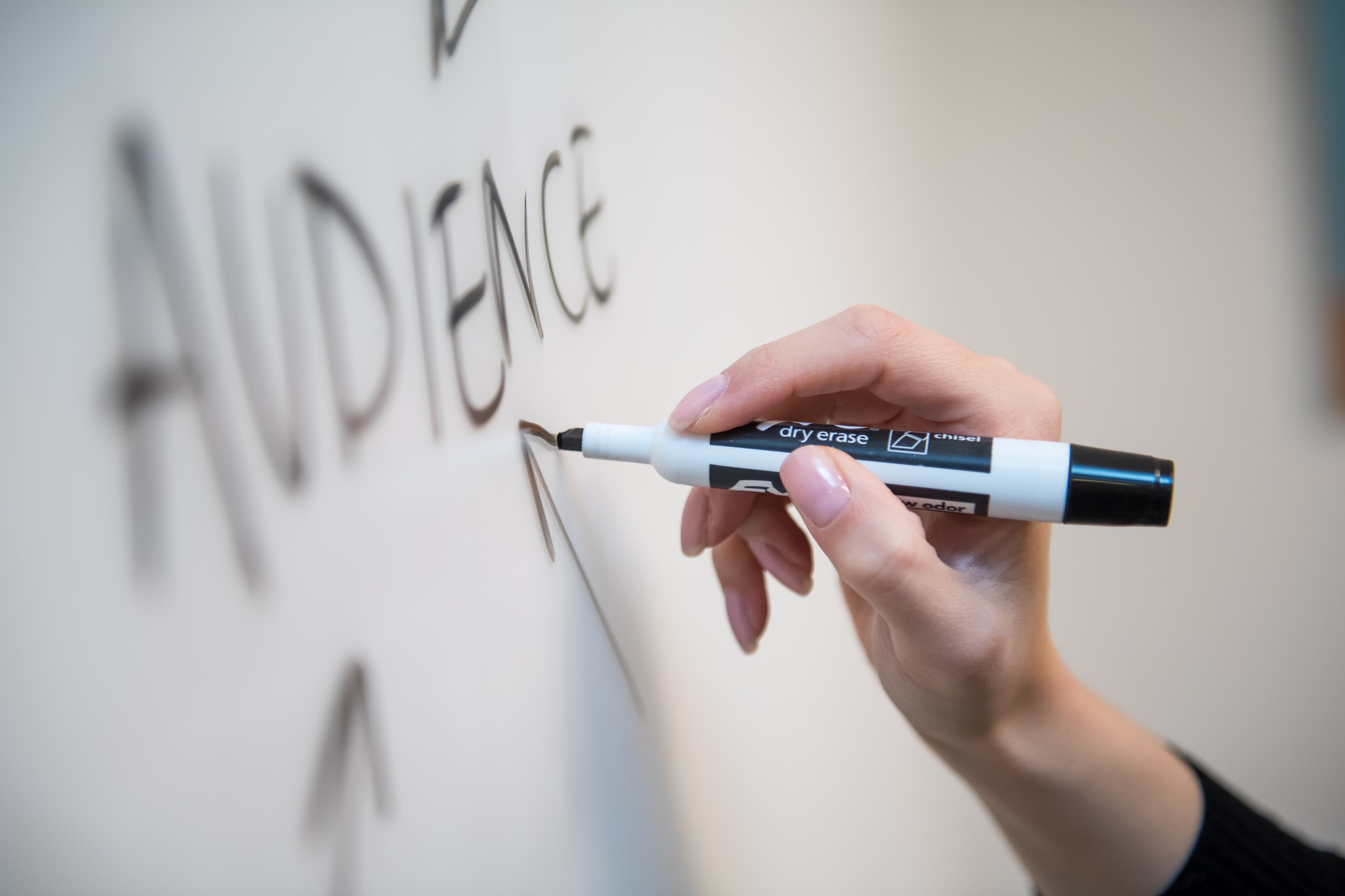 A hand writing the word Audience on a whiteboard with a marker pen.