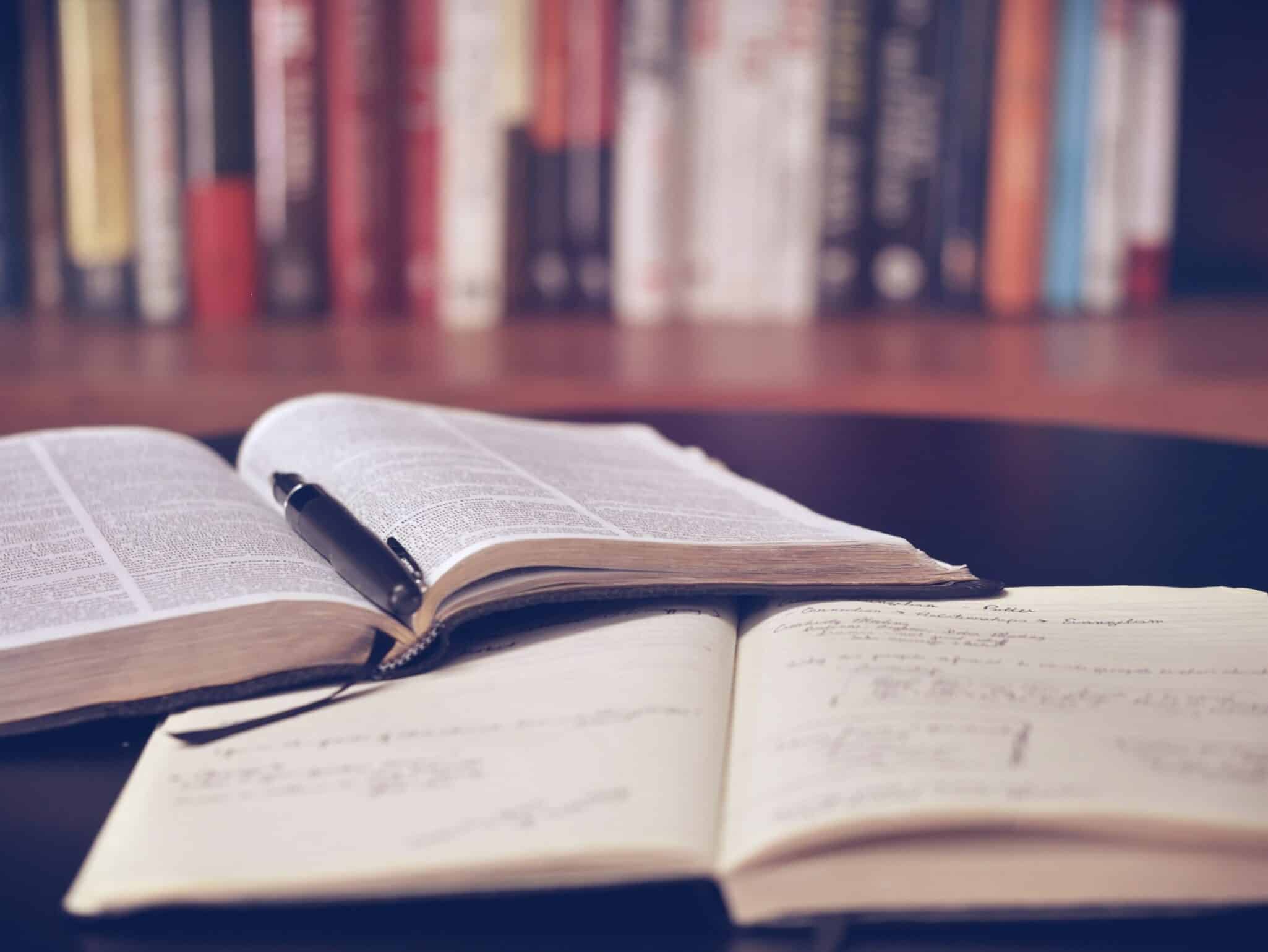 An open textbook and a handwritten notebook on a desk in front of a bookshelf.