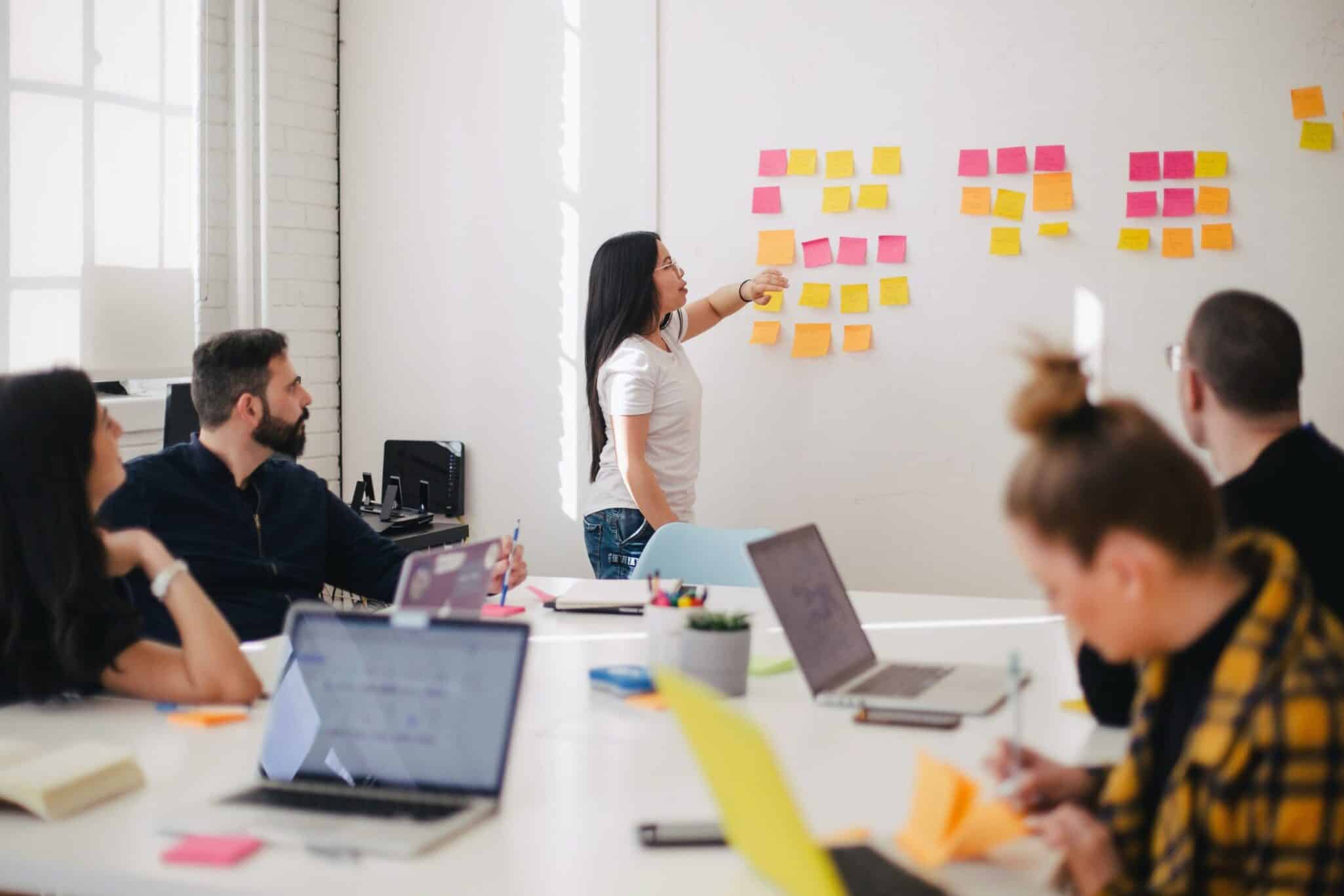 A woman leading a workshop, arranging sticky notes on a wall while colleagues watch from a meeting table.
