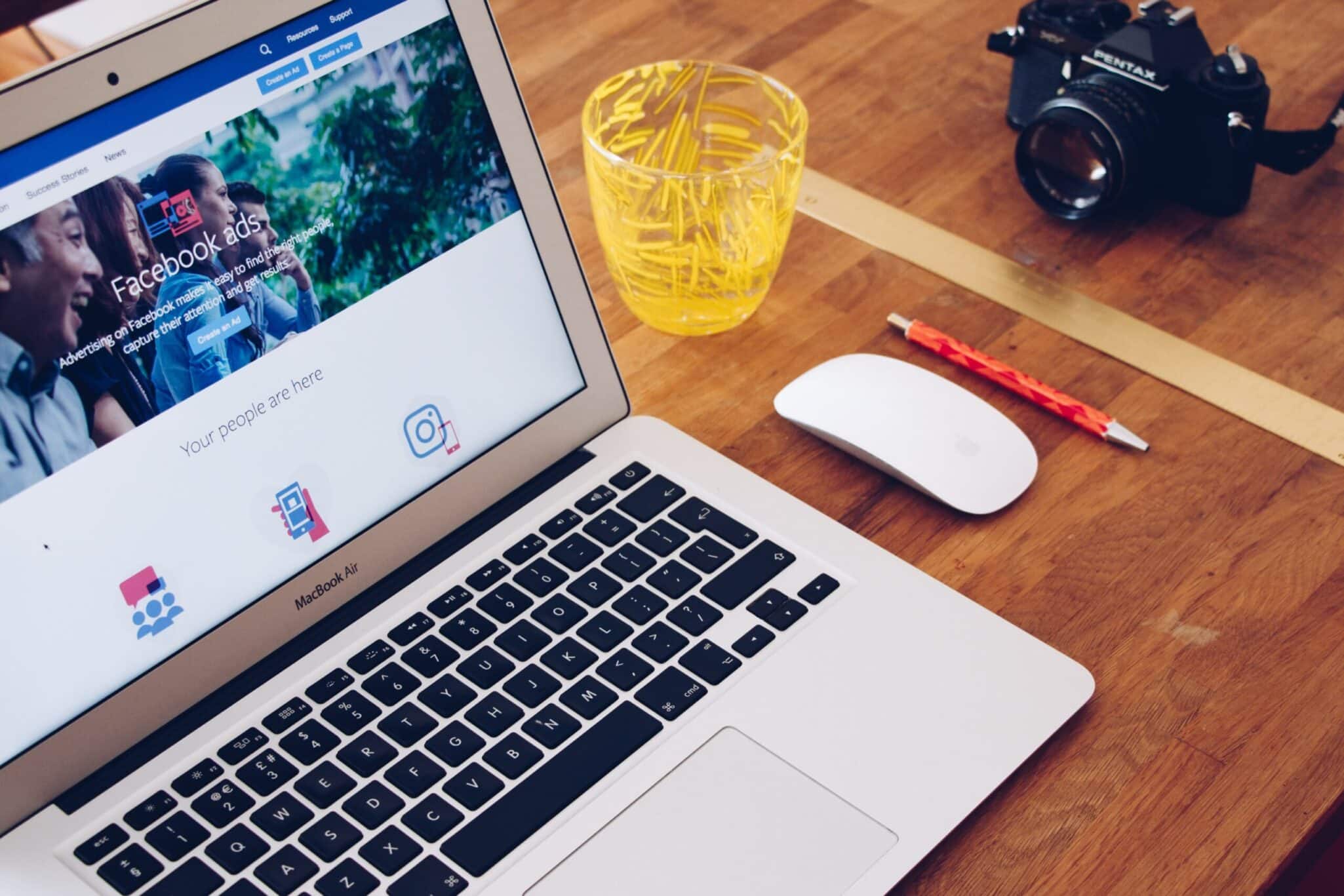A laptop showing a Facebook page on a wooden desk, beside a camera, a mouse and a pencil.