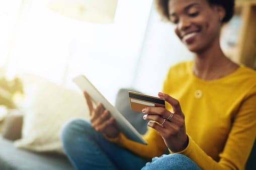 A woman sitting on a sofa holding a tablet in one hand and a bank card in the other.
