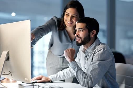 Two colleagues looking at a desktop monitor together, one pointing at the screen.