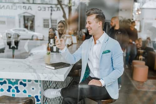 A man in a light blue jacket sitting at a cafe table, seen through the window.