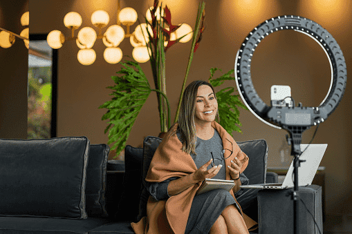 A woman recording herself on a phone with a ring light, sitting in a styled living room.