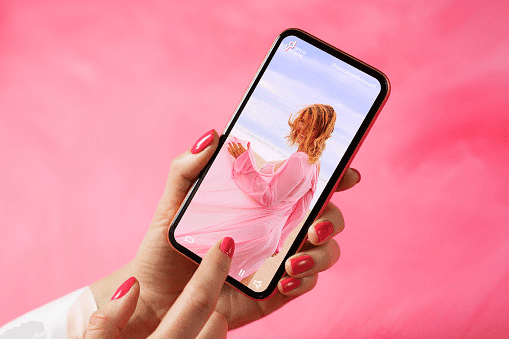 A hand holding a phone showing a fashion photo, against a pink feathered background.
