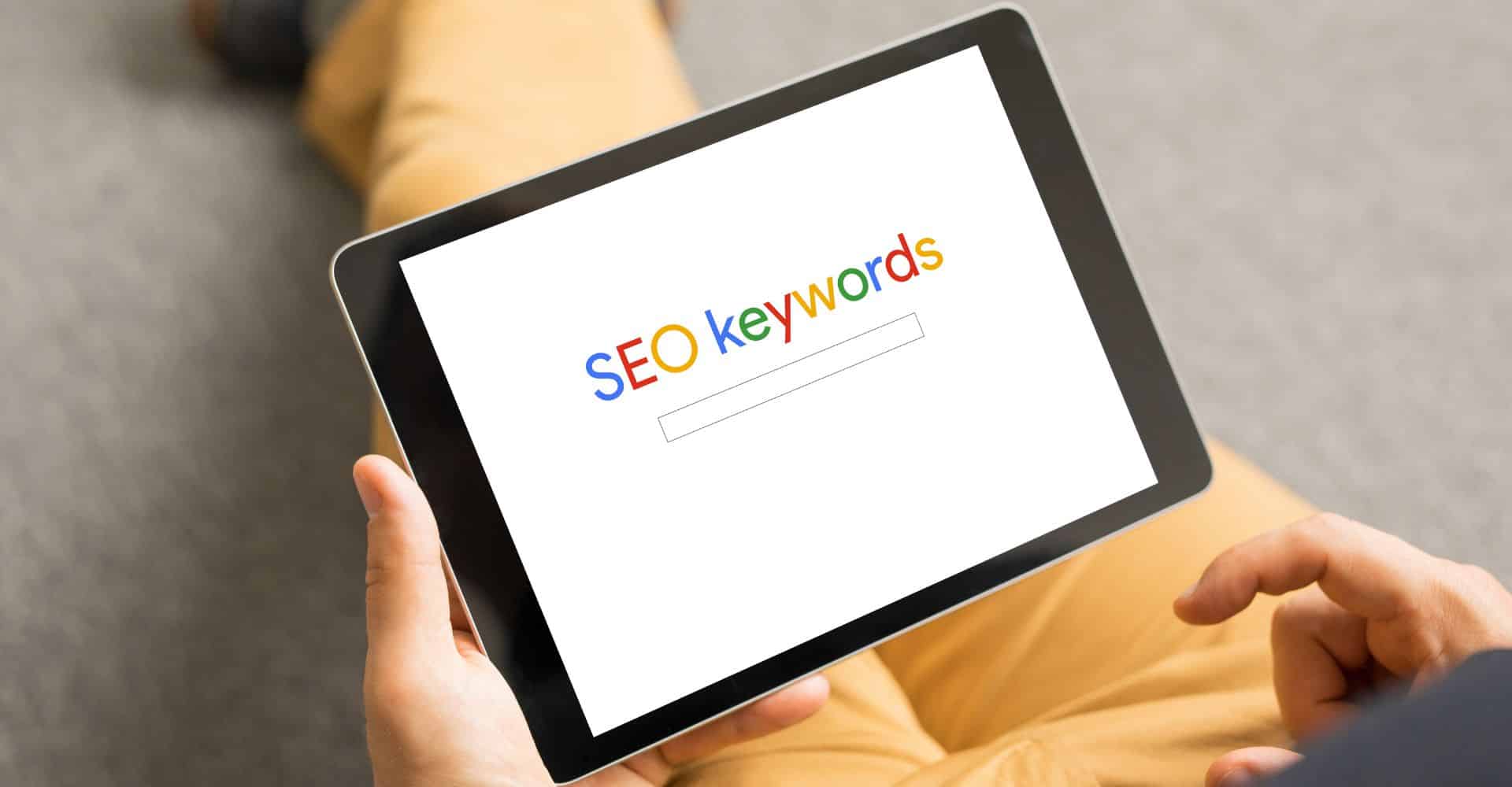 A tablet on someone's lap displaying the words SEO keywords in Google brand colours.