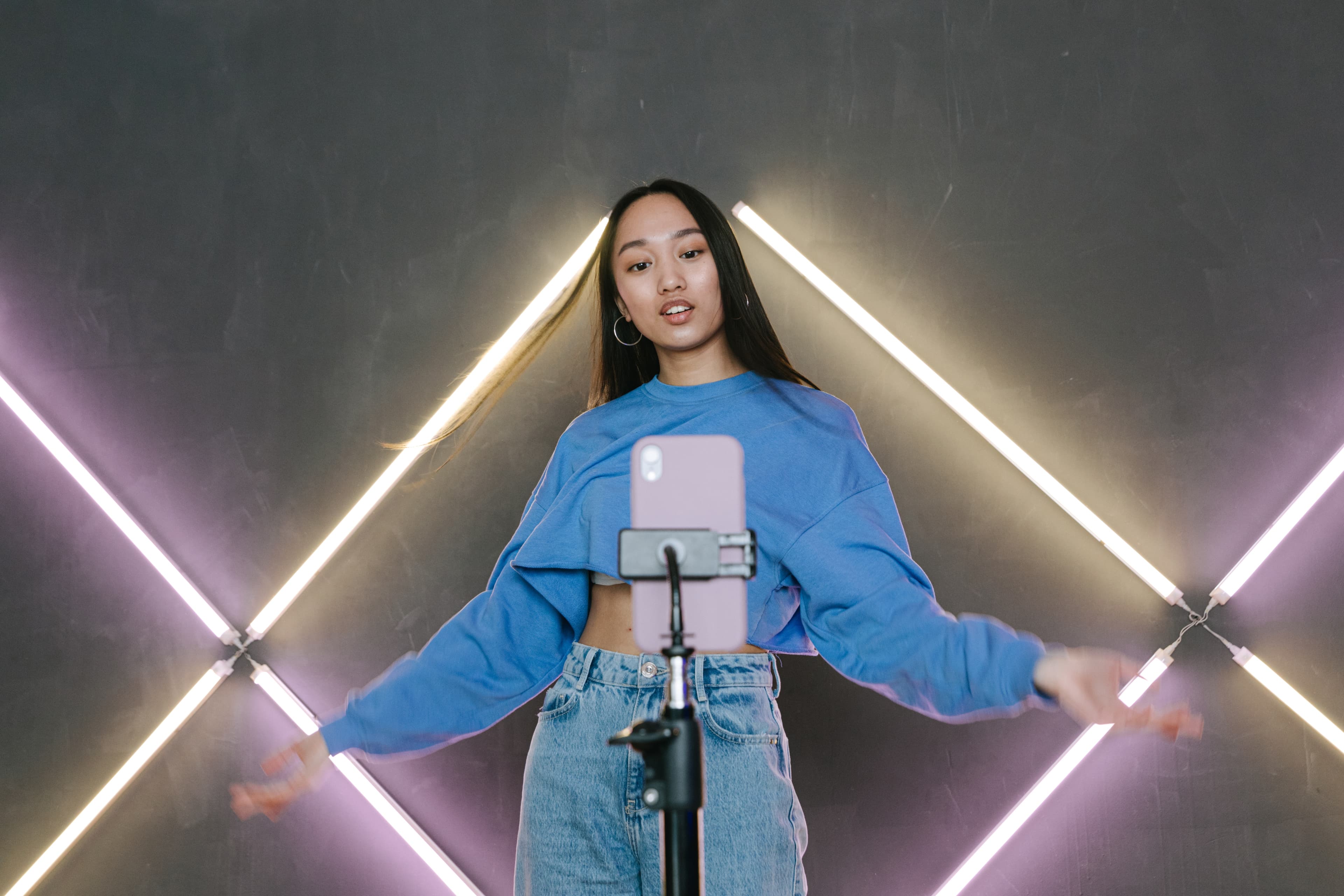 A woman recording a video on a phone mounted on a tripod, in front of a neon-lit wall.