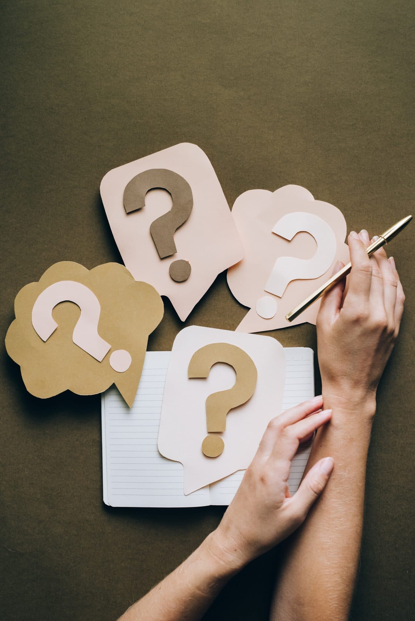 Paper speech bubbles with question marks on them, arranged on a notebook beside a pair of hands.