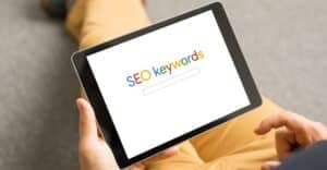 A tablet on someone's lap displaying the words SEO keywords in Google brand colours.