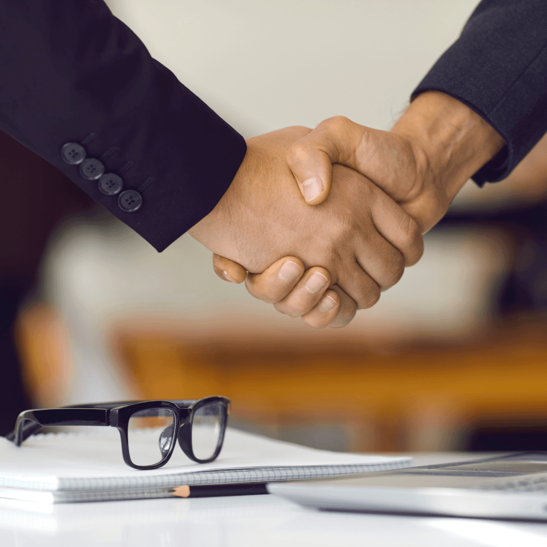 Two people in suits shaking hands over a desk with glasses and a notebook on it.