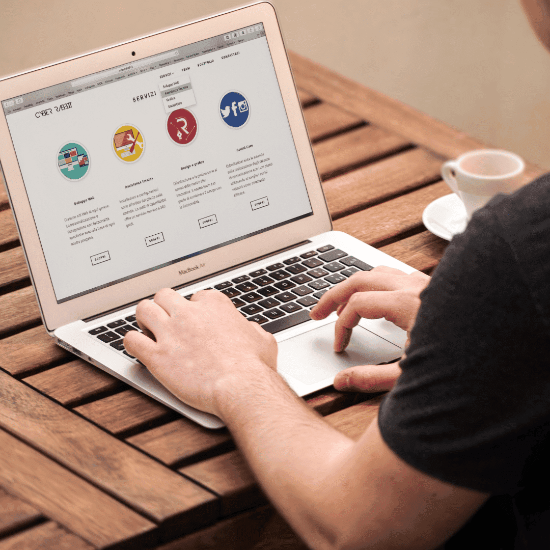 A man using a laptop at an outdoor wooden table, the screen showing a website with colourful service icons.