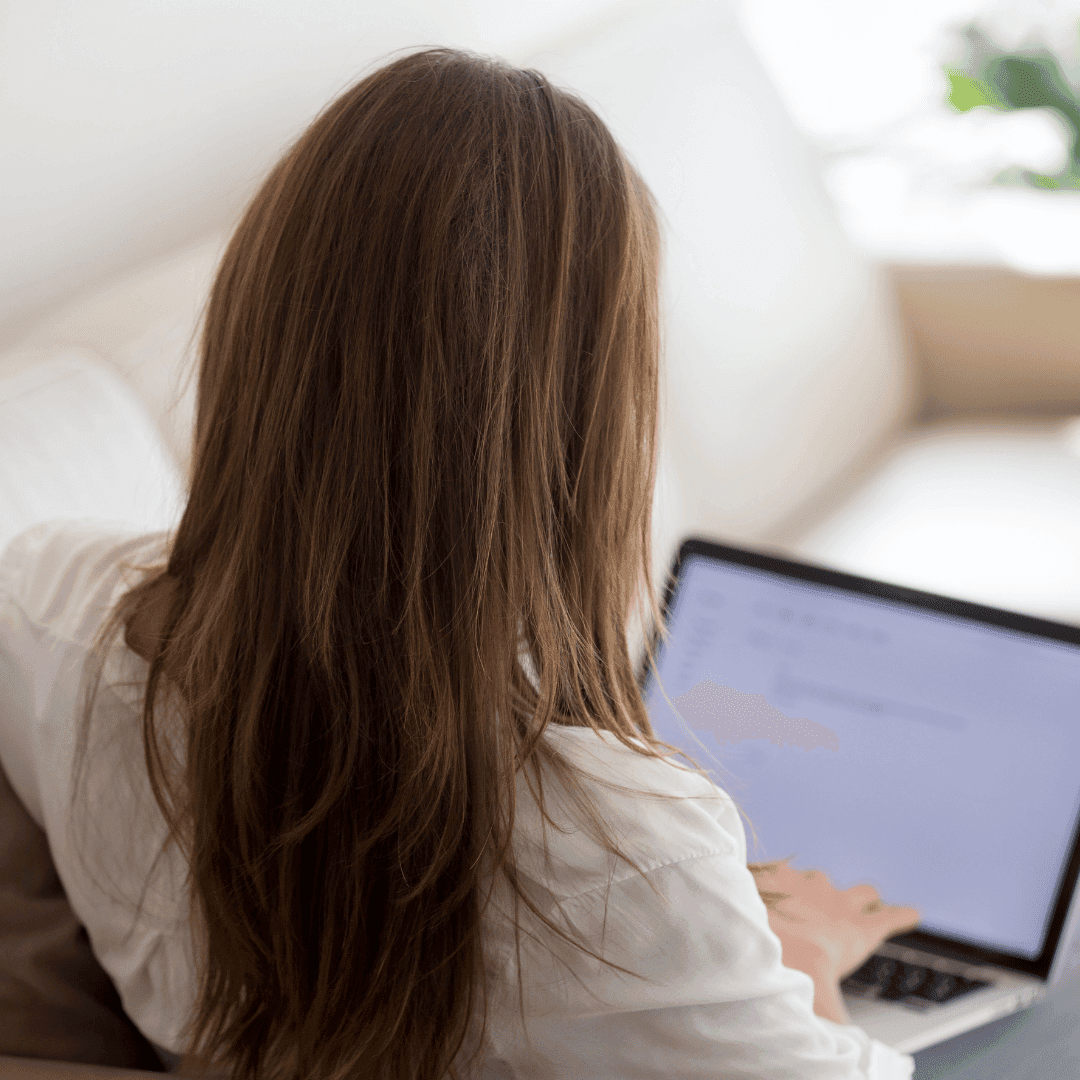 A woman sitting on a sofa working at a laptop, seen from behind.