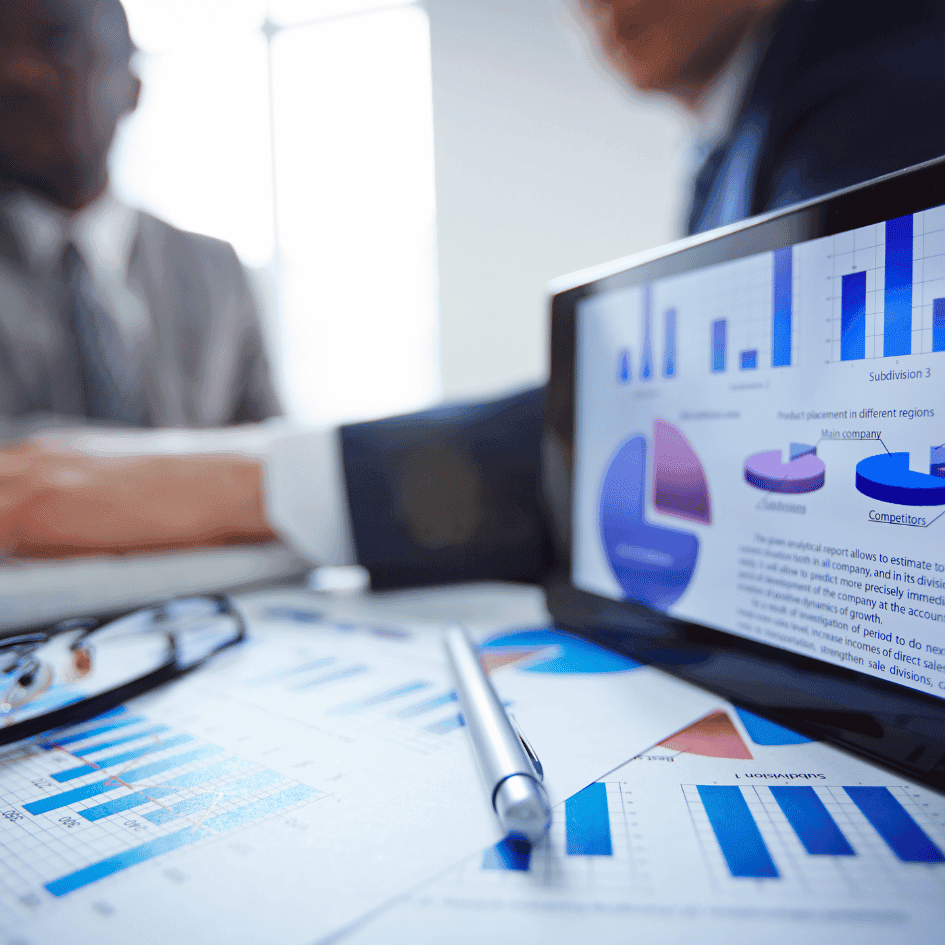 Two colleagues in a meeting behind a screen showing bar and pie charts, with printed reports on the desk.