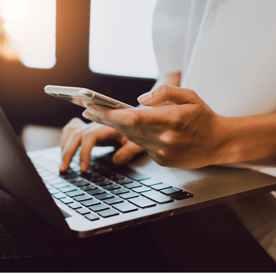 A woman holding a phone in one hand while typing on a laptop with the other.