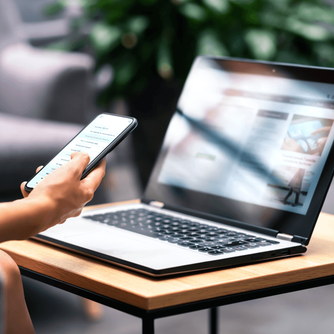 A person holding a phone beside an open laptop showing a web page.