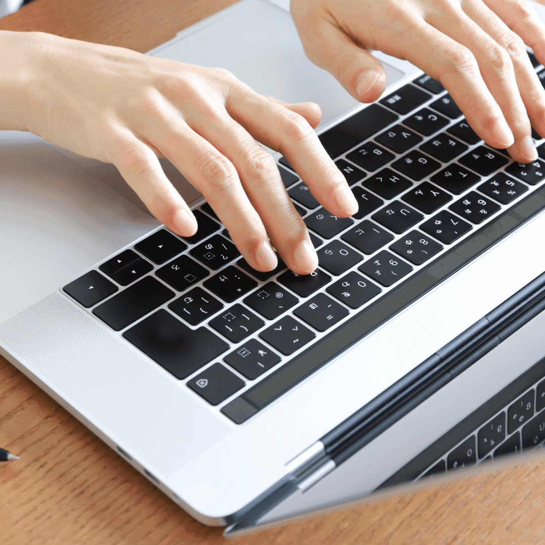 Close-up of two hands typing on a laptop keyboard at a wooden desk.