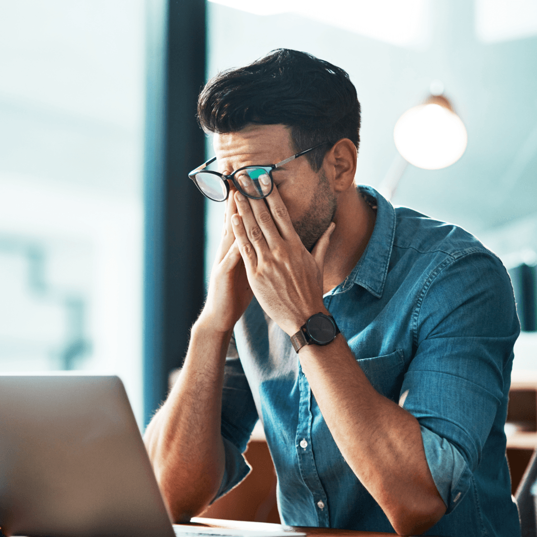A man at a laptop rubbing his eyes under his glasses, looking frustrated.