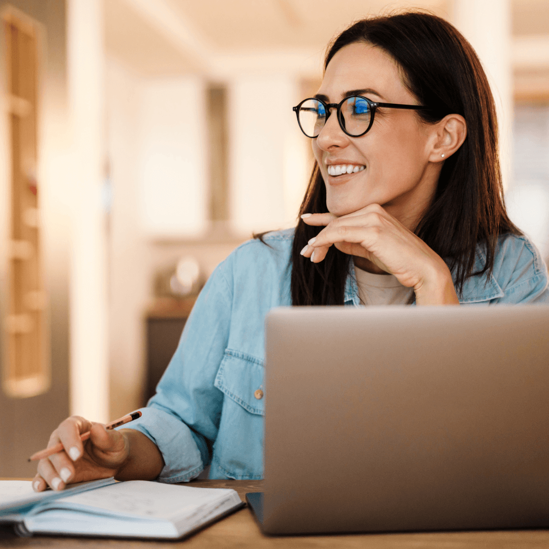 A woman in glasses smiling as she works at a laptop with a notebook beside her.