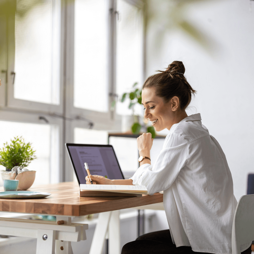 A woman making notes at a laptop in a bright home office.