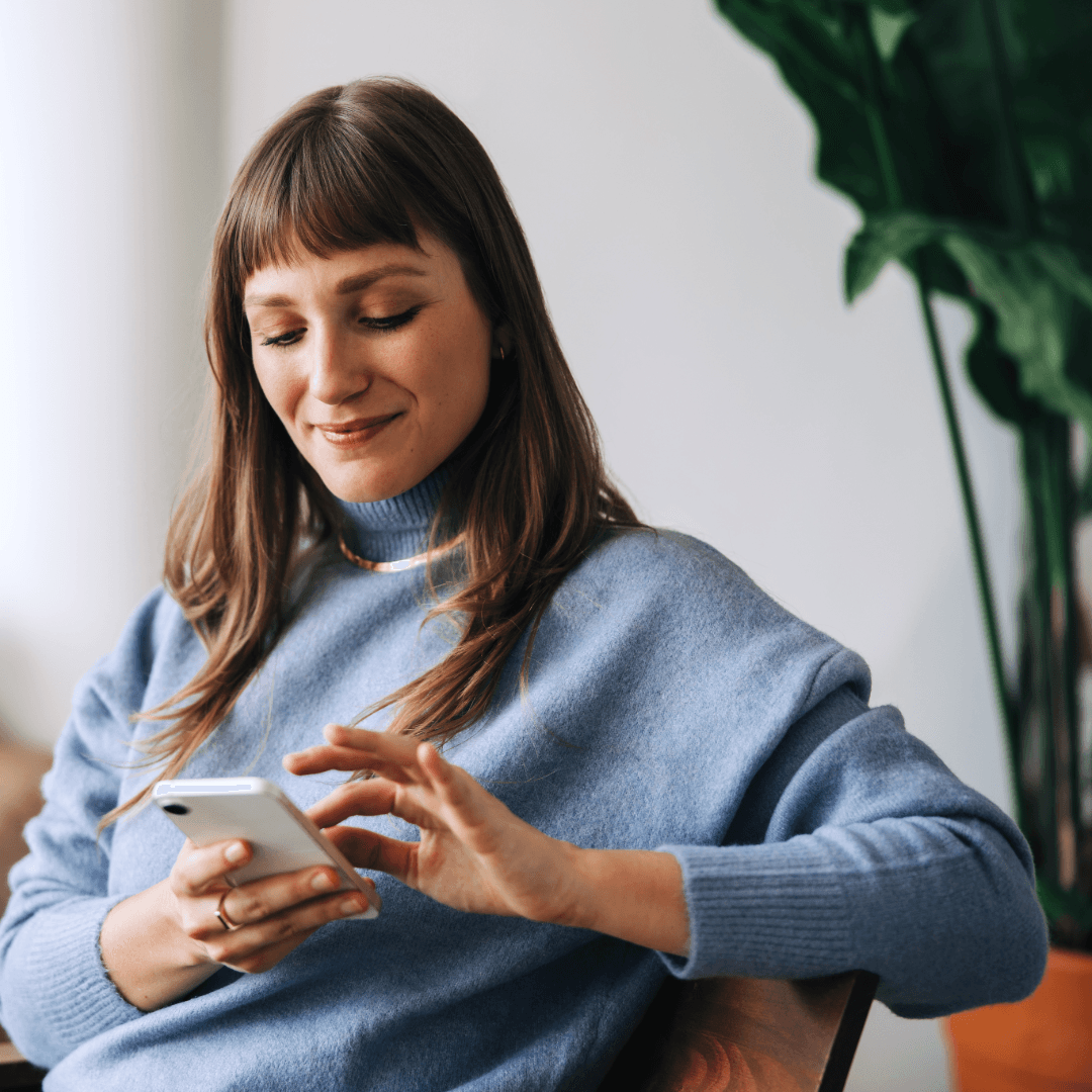 A woman sitting in an armchair smiling as she uses her phone.
