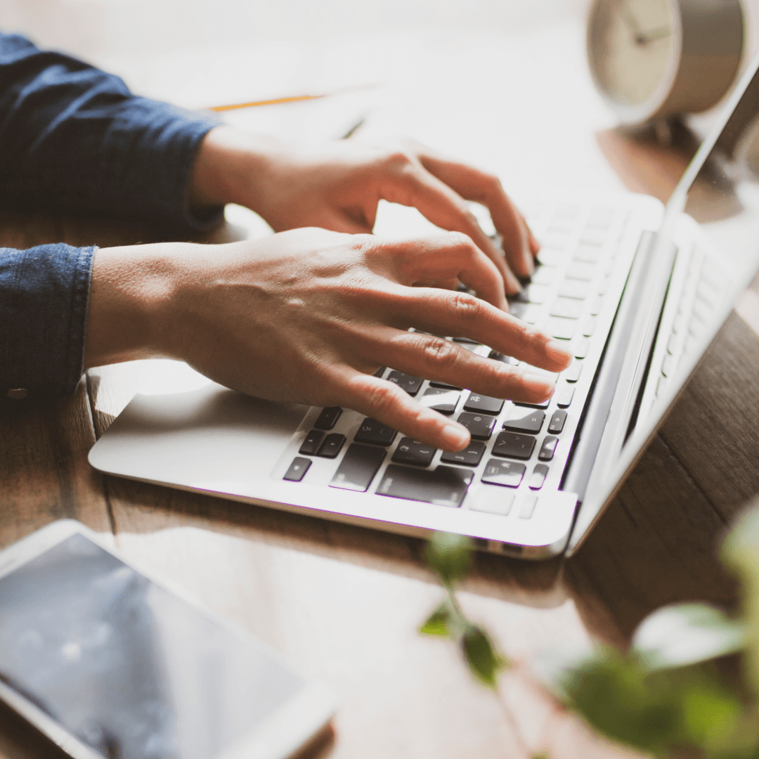 Hands typing on a laptop at a desk with a phone and a plant beside it.
