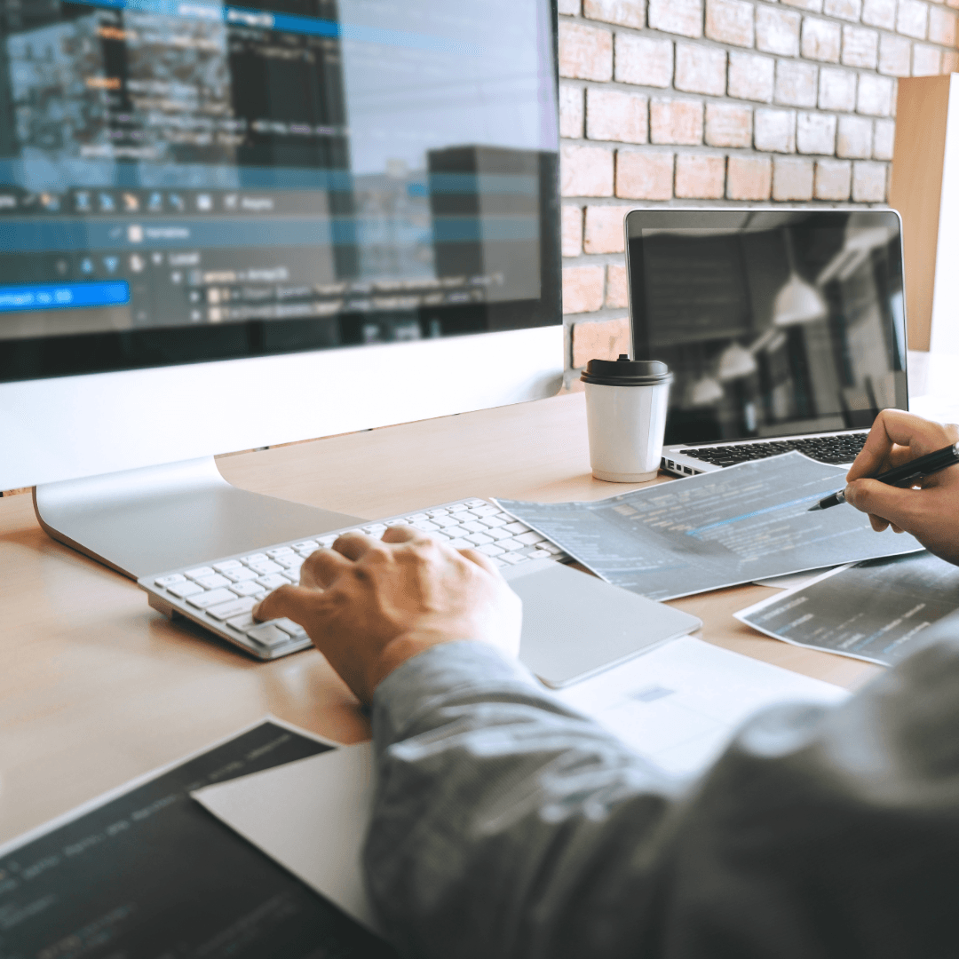 A web developer at a desk comparing code on a large monitor against printed page layouts.