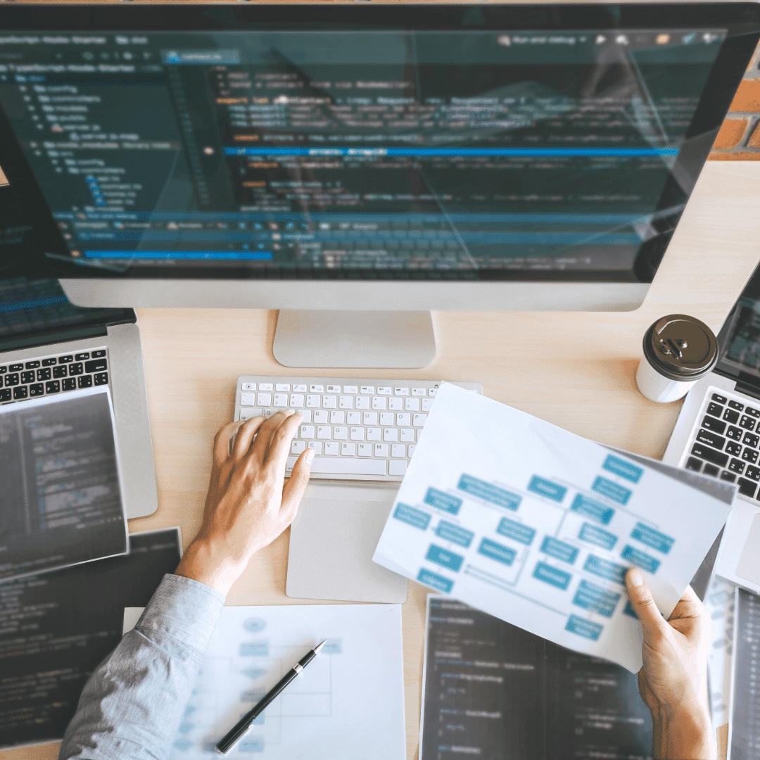 Overhead view of a developer at a desk, holding a printed site structure diagram beside a monitor full of code.
