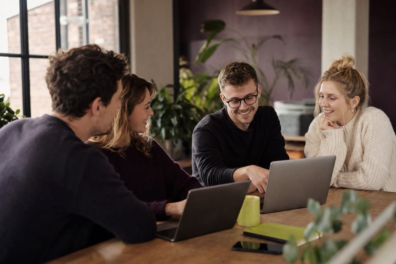 Four colleagues sitting around a wooden table with laptops, talking through a project together.