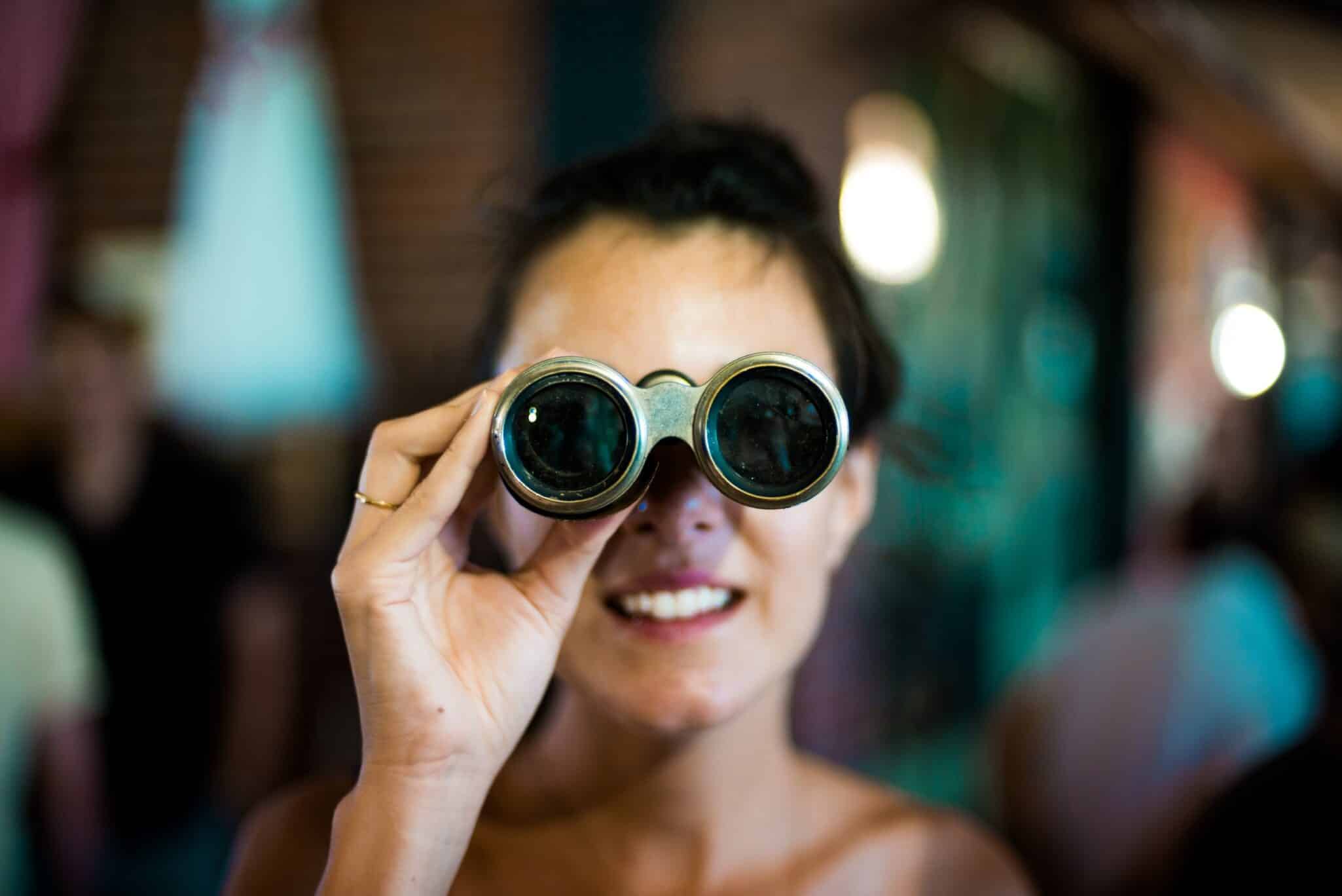 A smiling woman looking through a pair of small binoculars towards the camera.