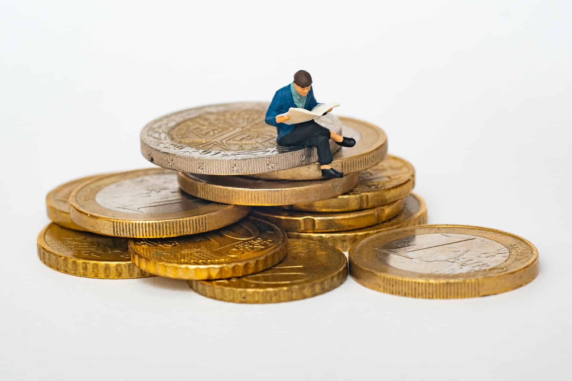 A miniature figure of a man reading a newspaper, sitting on a pile of euro coins.