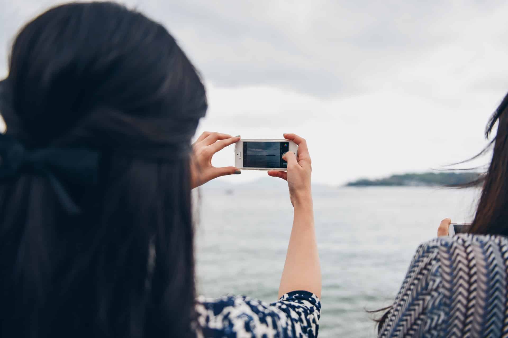 A woman photographing the sea on her phone, seen from behind.