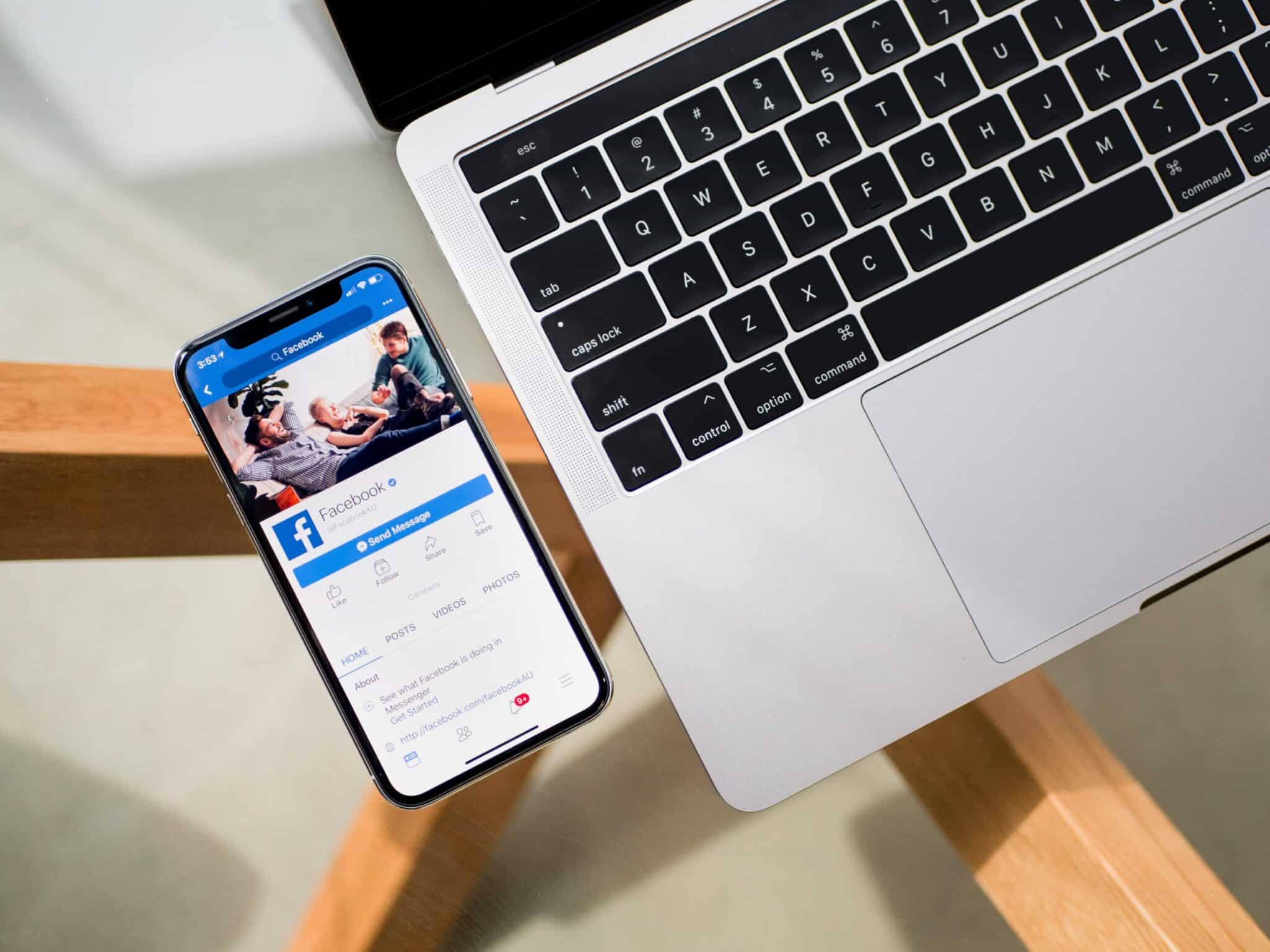 A phone showing a Facebook business page, lying next to an open laptop on a wooden desk.