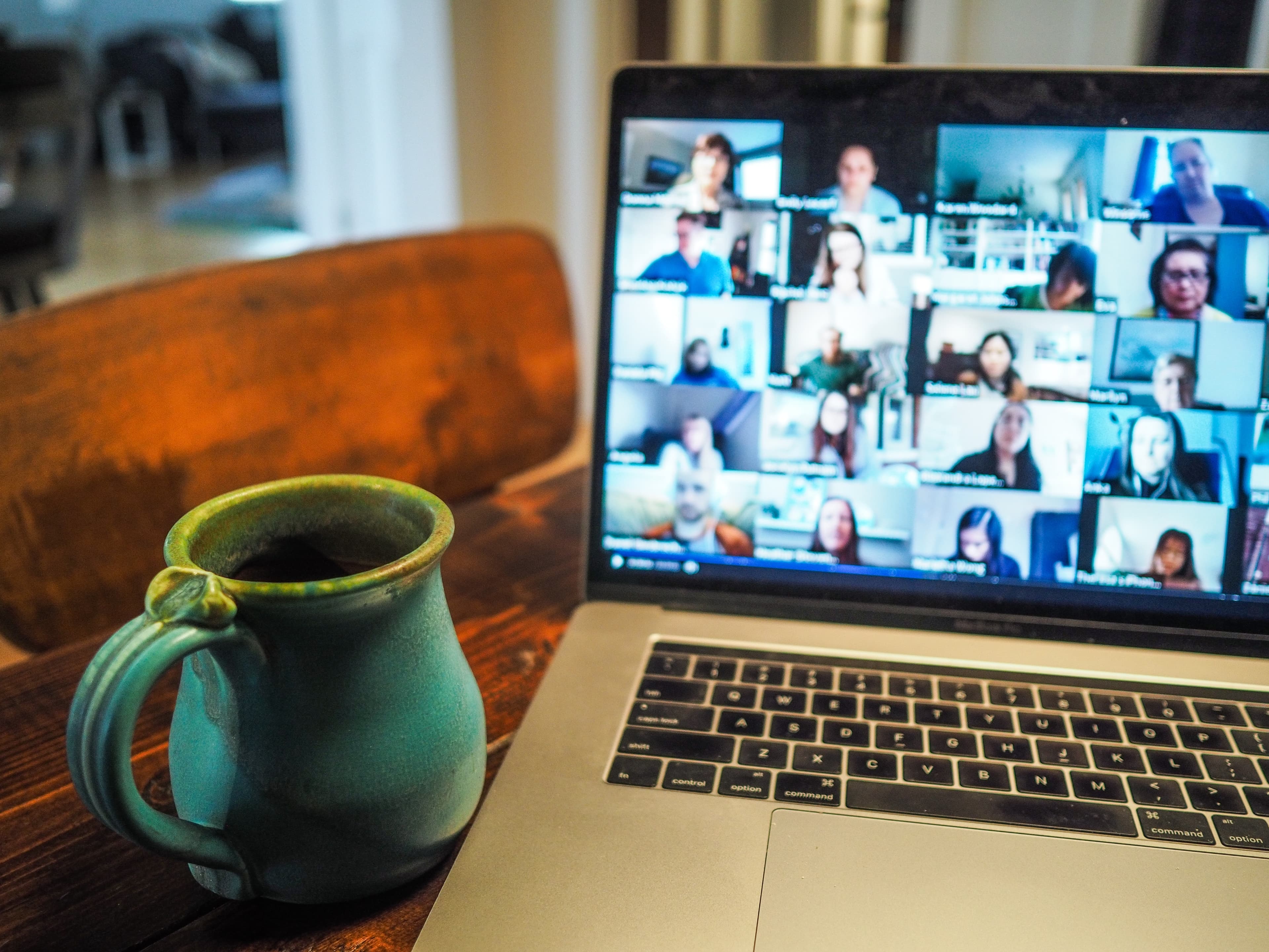 A laptop on a wooden table showing a grid of people on a video call, beside a mug of coffee.