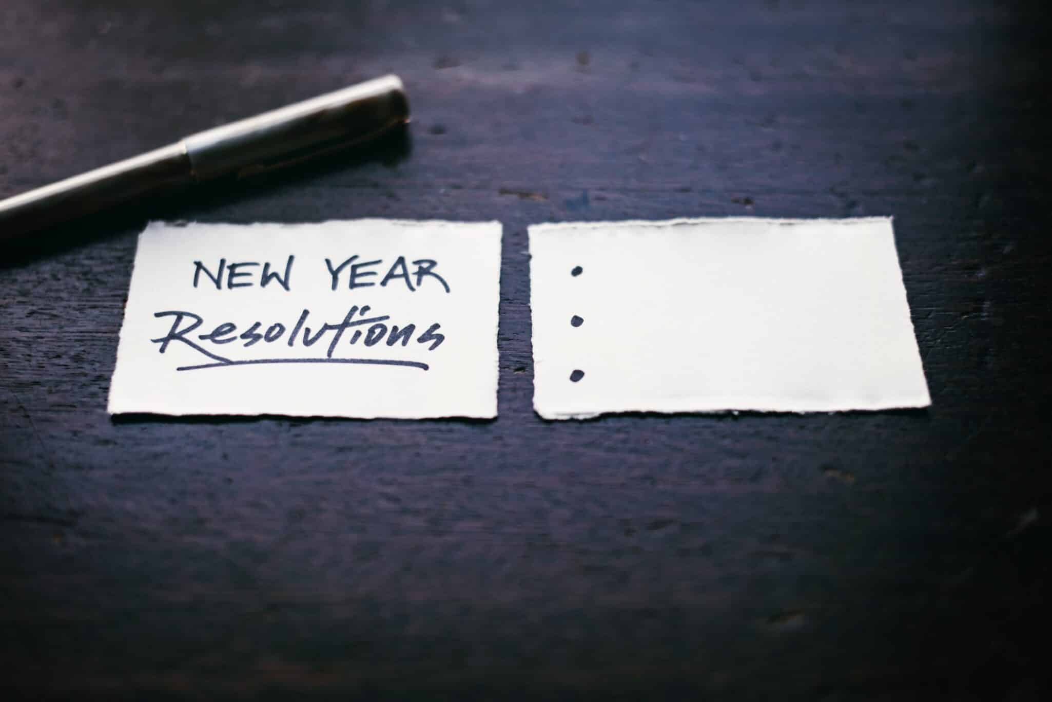 A torn piece of paper reading New Year Resolutions beside a blank list on a dark surface.