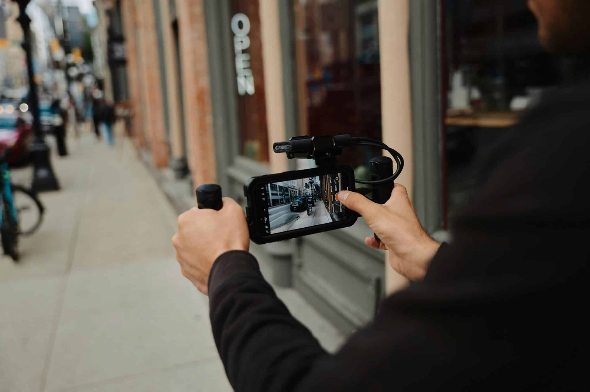 Over-the-shoulder shot of someone filming with a phone on a pathway beside a street.