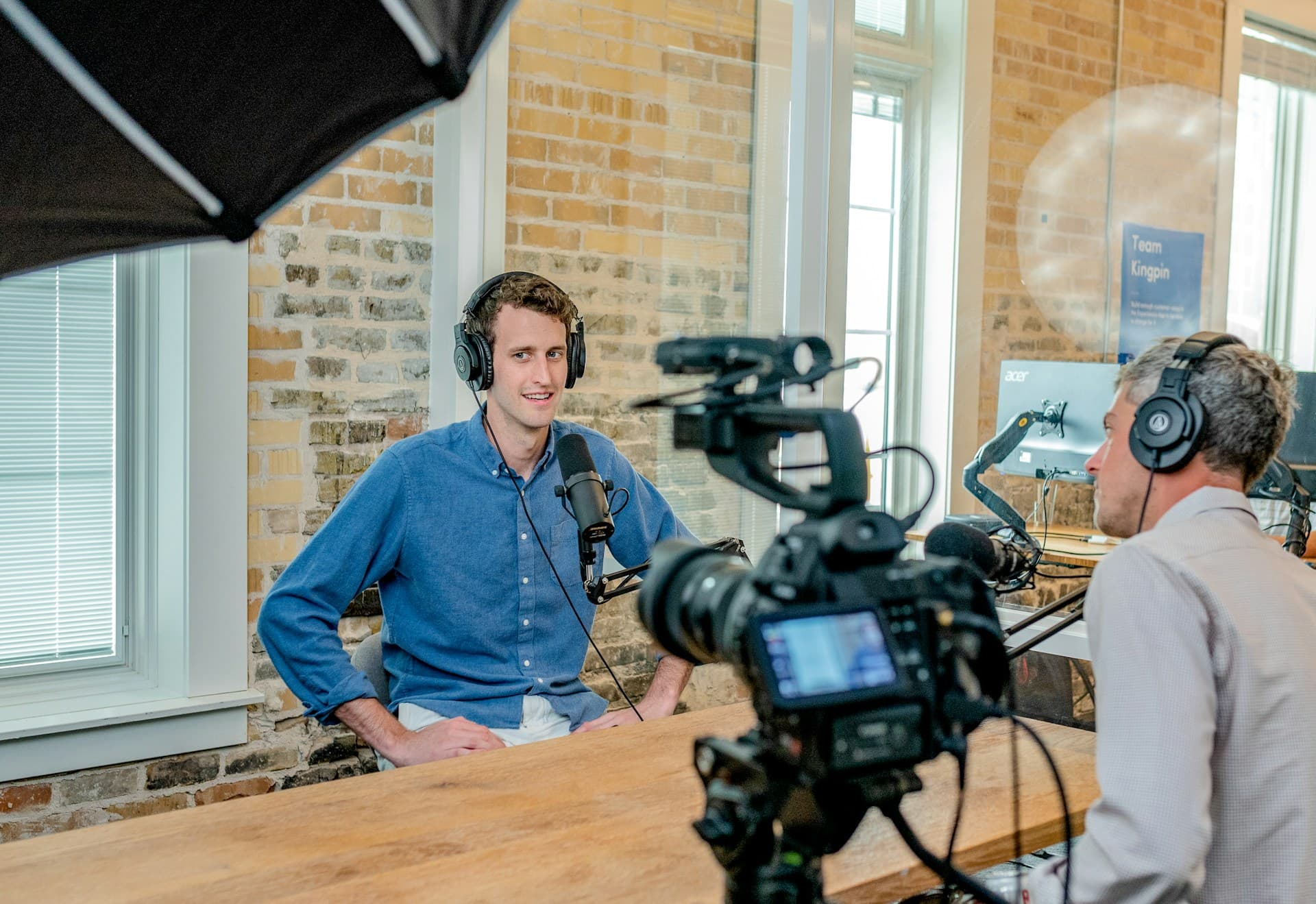 A man sits at a countertop facing the camera wearing headphones. A film crew member is filming him with a professional video camera. Storytelling in marketing.