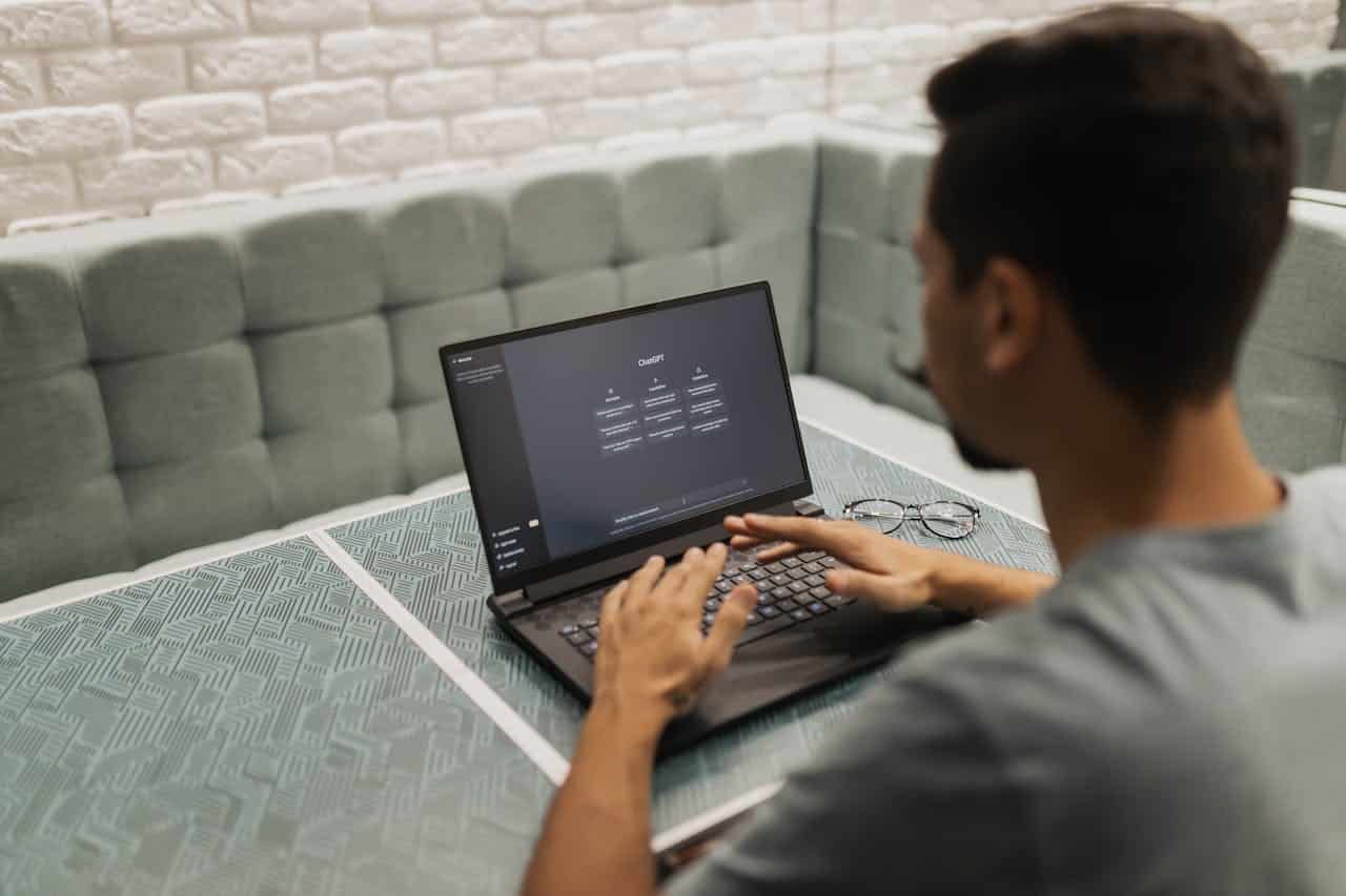 Image of a man sat at a cafe table using ChatGPT on his laptop. The photographer is looking over the man's shoulder. AI-powered content creation.